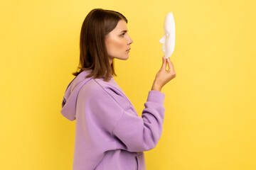 Profile of young adult attractive woman holding white mask in hands, hidden personality, expressing calm strict emotions, wearing purple hoodie. Indoor studio shot isolated on yellow background.