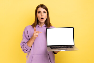 Portrait of surprised dark haired woman with big eyes pointing finger up and holding laptop with blank screen, having an idea, wearing purple hoodie. Indoor studio shot isolated on yellow background.