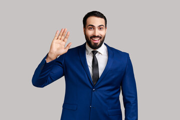 Satisfied handsome young bearded man standing waving hand, looking at camera with engaging toothy smile, wearing official style suit. Indoor studio shot isolated on gray background.