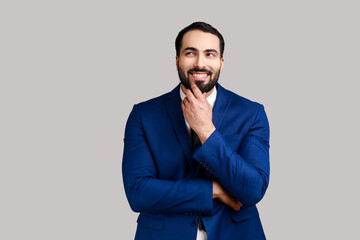 Bearded man with peaceful face thinking over plans for future holding his chin, thoughtful male dreaming about good job, wearing official style suit. Indoor studio shot isolated on gray background.