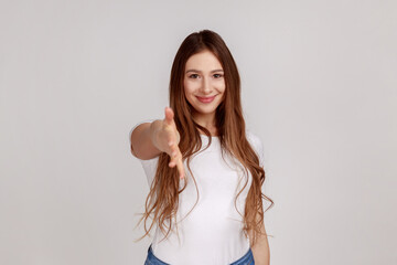 Let me introduce myself. Portrait of friendly woman giving hand to handshake, greeting guests with toothy smile, wearing white T-shirt. Indoor studio shot isolated on gray background. © khosrork