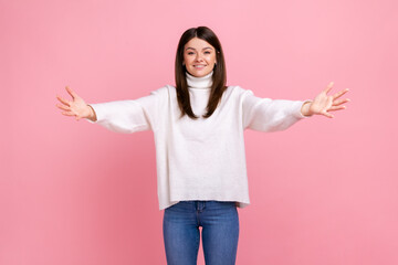 Portrait of happy friendly woman outstretching hands to embrace, giving free hugs and welcoming, wearing white casual style sweater. Indoor studio shot isolated on pink background.