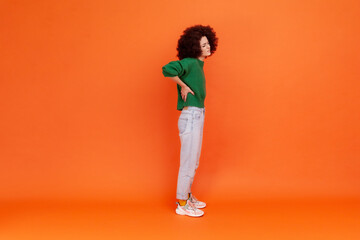 Fototapeta na wymiar Full length portrait of woman with Afro hairstyle wearing green casual style sweater touching pain lower back, having problems with kidney. Indoor studio shot isolated on orange background.