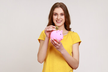 Portrait of young adorable female putting physical bitcoin into piggy bank, investment in cryptocurrency concept, looking smiling at camera. Indoor studio shot isolated on gray background.