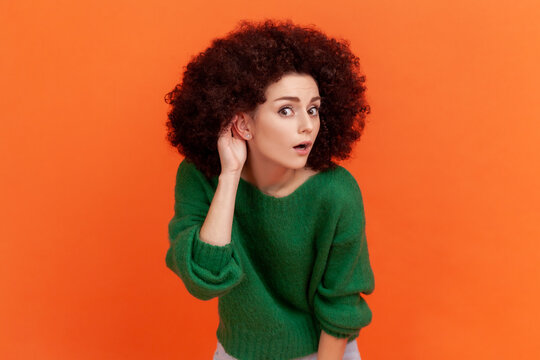 Woman with Afro hairstyle wearing green casual style sweater keeping hand near ear to listen better, having hearing problems, difficult to understand. Indoor studio shot isolated on orange background.