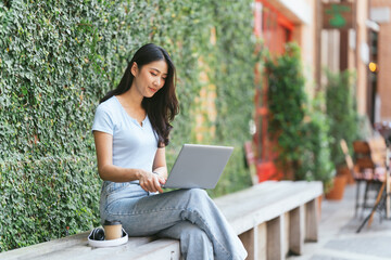 Portrait of beautiful Asian woman sitting outdoors at coffee shop restaurant during summer, using smart wireless technology computer laptop and smartphone, relaxing coffee break at cafe restaurant.