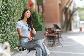 Portrait of beautiful Asian woman sitting outdoors at coffee shop restaurant during summer, using smart wireless technology computer laptop and smartphone, relaxing coffee break at cafe restaurant.