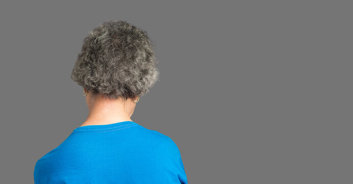 Back view of a senior woman suffering neck pain and neck tilt while standing against a gray background - Powered by Adobe