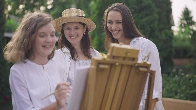 Curios pregnant woman and young friend looking at blurred teenage girl painting in slow motion on easel. Portrait of smiling Caucasian women admiring talented teen enjoying hobby on spring day