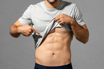 Muscular male model lifting up t-shirt to show abs while standing on gray isolated background in...