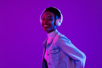 Fototapeta Stylish happy smiling African-American woman wearing headphones and listening to music in modern purple studio background obraz