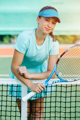 Awkward smiling girl leaning on a tennis court net in a light blue sportswear