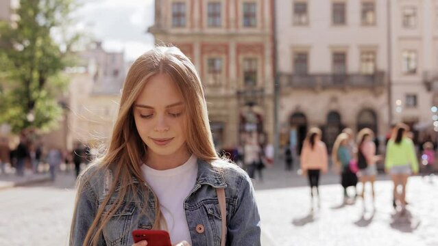 Attractive young blond woman use cellphone smiling. Walking on street in city center. Travel tourist cellphone outdoors. Portrait. Close up of joyful smiling woman tapping on mobile phone outdoors.
