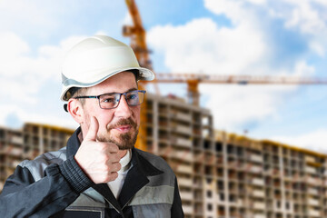 A male builder in a white hard hat against a blurred background of a construction site with a blue sky. Positive civil engineer with a beard.