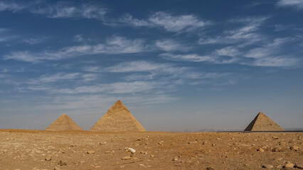 The three great pyramids of Giza - Cheops, Khafre, Menkaur - against the blue sky and clouds. In the foreground is a rocky-sandy desert. Silhouettes of Cairo buildings on the horizon. Egypt