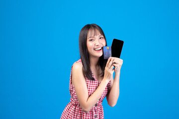 Portrait Asian happy young girl smiling cheerful and showing plastic credit card while holding mobile phone isolated on blue background