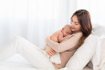 Asian young mother holding tenderly cuddles newborn baby in arms on to chest. Mother looking adorable infant with love and care. Woman and small child lying on bed together at home.