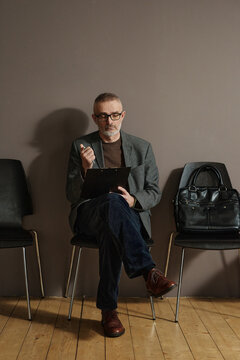 Mature man in eyeglasses sitting on chair in hallway and and filling the form of resume before job interview
