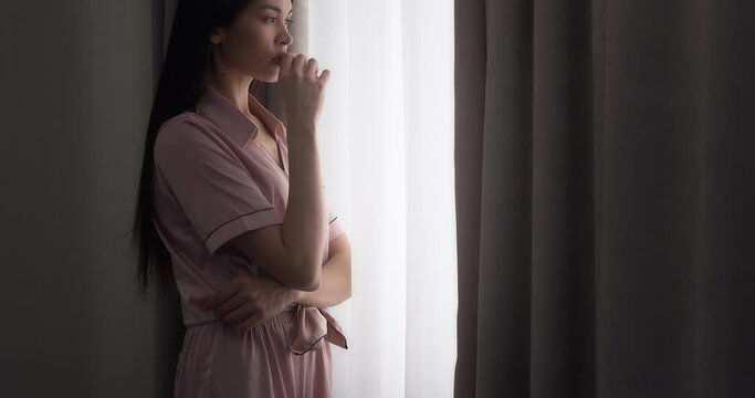 Upwards. Young woman with long dark hair dressed in pink silk pajamas is standing near the window in a room at home and vaping, smoking an eron cigarette, blowing smoke.