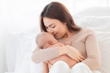 Selective focus Beautiful Asian mother holding newborn baby and kiss baby with love and tender. Adorable infant deepy sleeping on her chest. Woman and toddler spend time together at home.