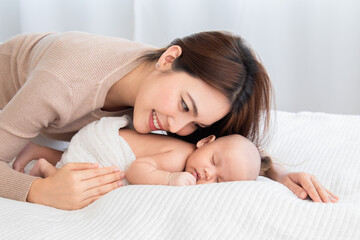 Beautiful asian mom supports and tenderly cuddles newborn baby gently while baby deepy sleeping on bed. Young woman looking adorable infant with love and tender. Mother and child spend time together.