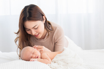 Beautiful asian mom supports and tenderly cuddles newborn baby gently while baby deepy sleeping on bed. Young woman looking adorable infant with love and tender. Mother and child spend time together.