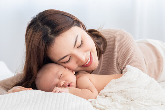 Beautiful asian mom supports and tenderly cuddles newborn baby gently while baby sleeping. Young woman kissing and touching on back child with love and tender. Mother and infant spend time together.