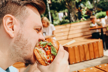Young hungry caucasian man eating hamburger or beef burger with vegetables and sauce in outdoor...