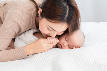 Fototapeta na wymiar Selective focus beautiful Asian mother kiss hand newborm baby with love and care. Woman hold tiny finger adorable infant while sleeping comfortable on bed at home. Mom and toddler spend time together.