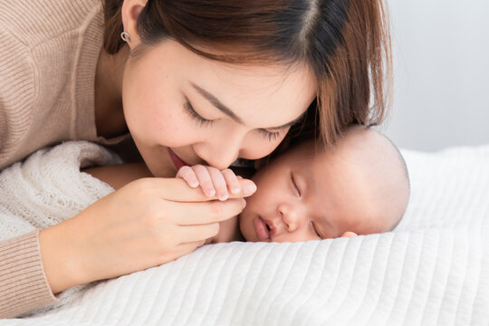 Selective focus beautiful Asian mother kiss hand newborm baby with love and care. Woman hold tiny finger adorable infant while sleeping comfortable on bed at home. Mom and toddler spend time together.