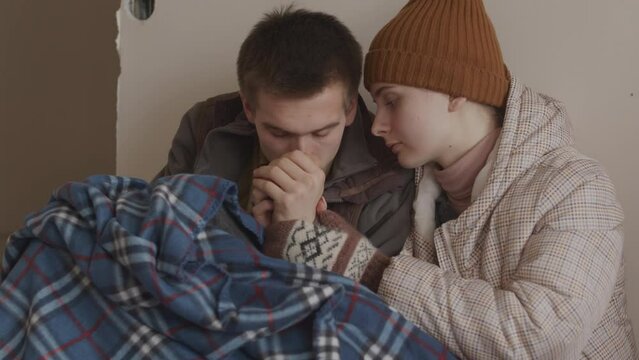 Medium slowmo of young Caucasian couple of refugees warming up each others hands sitting close to each other under old checkered plaid in asylum