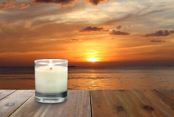 the burning luxury aromatic scented candle glass on the wooden table with background of sea beach during sunset at the evening