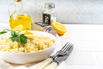Cooked couscous with cilantro or parsley , butter, olive oil, lemon in white ceramic bowl on kitchen white table copy space