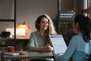 Young woman sitting at table and smiling while manager examining her resume during job interview - obrazy, fototapety, plakaty