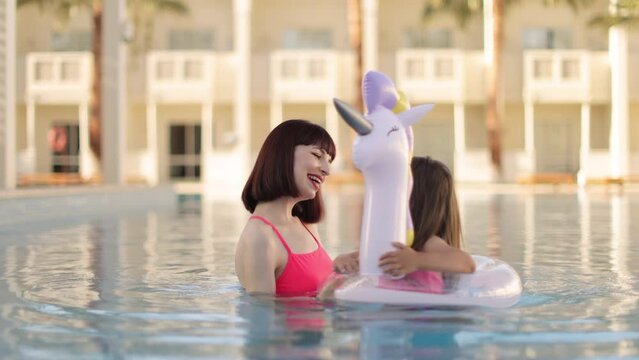 Mom plays with her little cute daughter with unicorn inflatable ring in the pool against the background of a summer exotic resort. Mother and daughter enjoying vacation by the pool