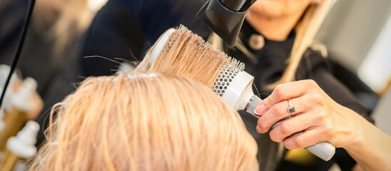 Drying straight blond hair with black hairdryer and white round brush in hairdresser salon, close up