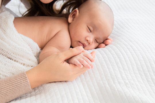Selective focus of newborn baby deepy sleeping on white bed. Asian mother supports and holding hand adorable infant gently while toddler sleeping on bed. Child sleep in mom's hand.
