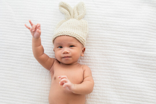 Asian adorable newborn baby wearing pants and rabbit hat beige knitted lying on white bed. little baby 0-1month looking surprised at camera. concept of happy childhood and motherhood.