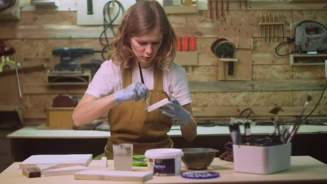 A woman paints a wooden part of a wooden product with a brush. Business idea for a home workshop