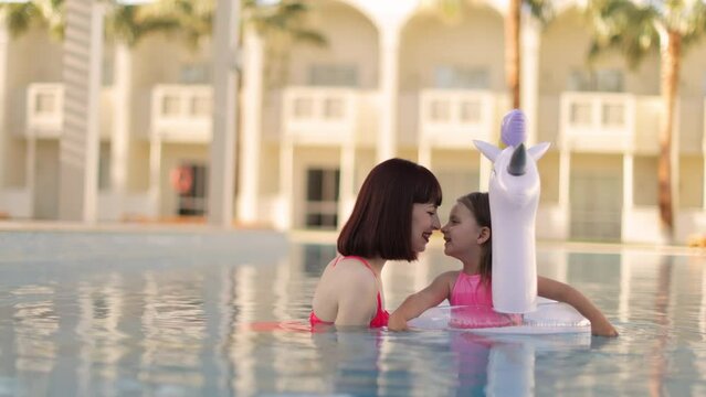 Mom plays with her little cute daughter with unicorn inflatable ring in the pool against the background of a summer exotic resort. Mother and daughter enjoying vacation by the pool