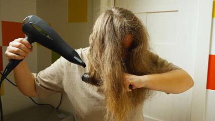 Close-up of a woman in the bathroom drying her hair with an electric hair dryer and comb. Everyday preparations before work.