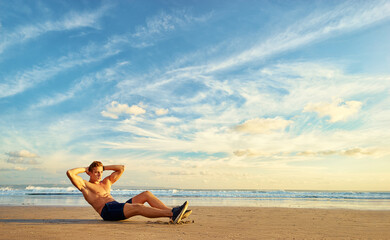 Sports and healthy lifestyle. Young man doing crunches on the ocean beach. Enjoying sunset.