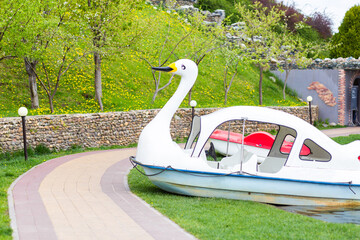 A catamaran in the form of a white swan stands on land near a clear lake on a bright sunny day.