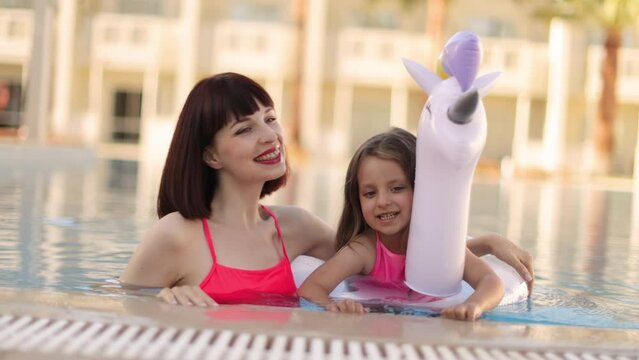 Young girl kid with unicorn inflatable ring splashing in swimming pool having fun leisure activity. Happy mother and her daughter in the same pink swimsuits playing in swimming pool. Summer vacations