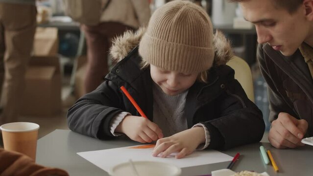 Waist up shot of 7 year old Caucasian refugee boy in hat and warm outwear drawing while sitting at table in local food bank with other refugees
