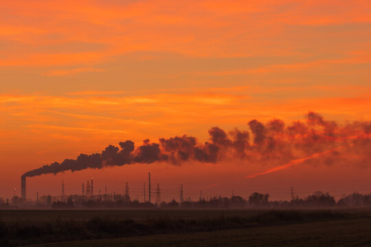a long tail of smoke streaming across the sky from a refinery against a red sky during sunset