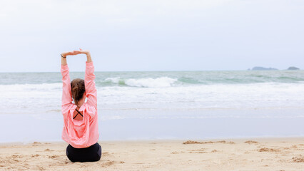 asian woman wearing sportwear and pink jacket sitting exercise and relax on the sea beach at evening in the rain season