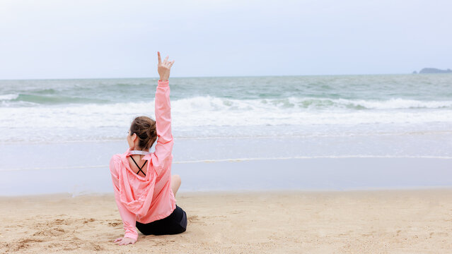 asian woman wearing sportwear and pink jacket sitting exercise and relax on the sea beach at evening in the rain season