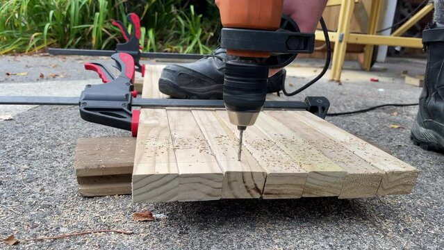A man uses a power tool to pre-drill a row of holes into pieces of timber clamped together on the ground