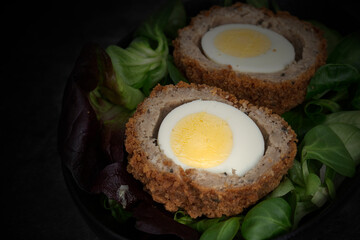 Scotch egg halved on lettuce leaves in a black dish.  On a stone background